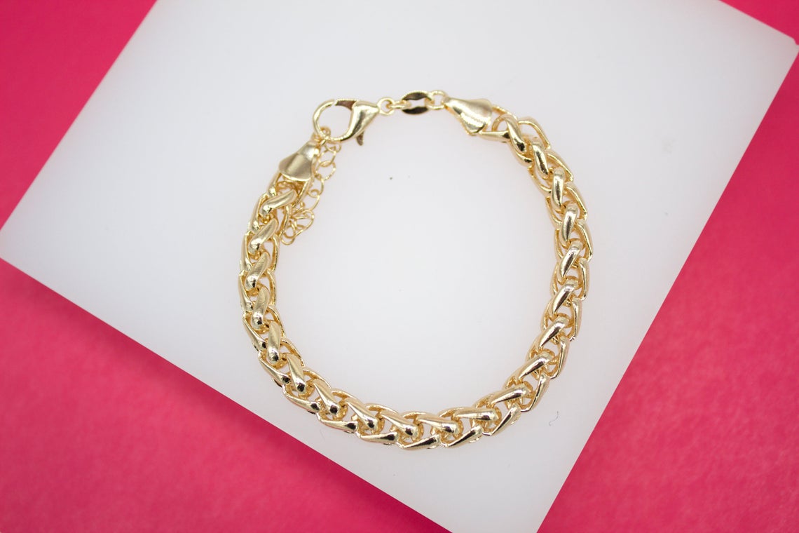 A thick gold link bracelet rests on a white display stand against a vibrant pink background.