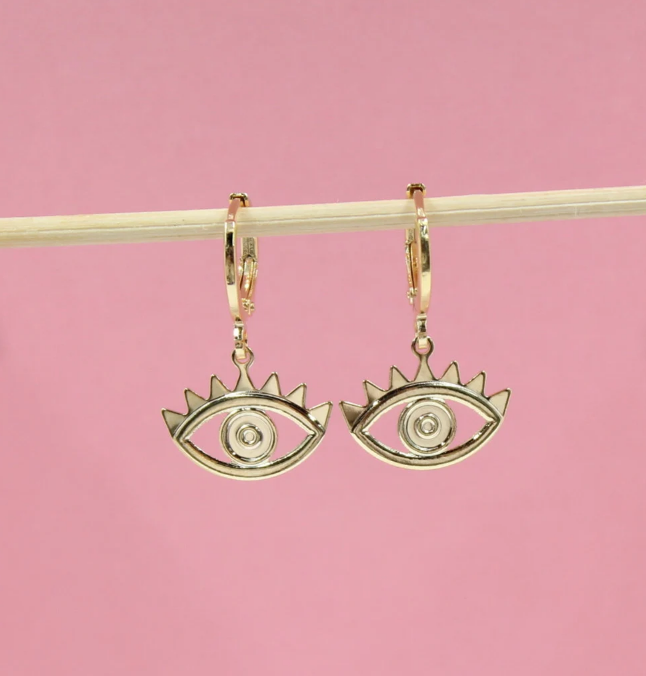 Two gold evil eye hoop earrings hang from a wooden stick against a soft pink background.
