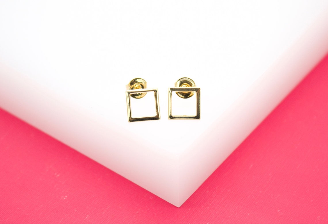 A pair of geometric square gold stud earrings rests elegantly on a white and pink display.