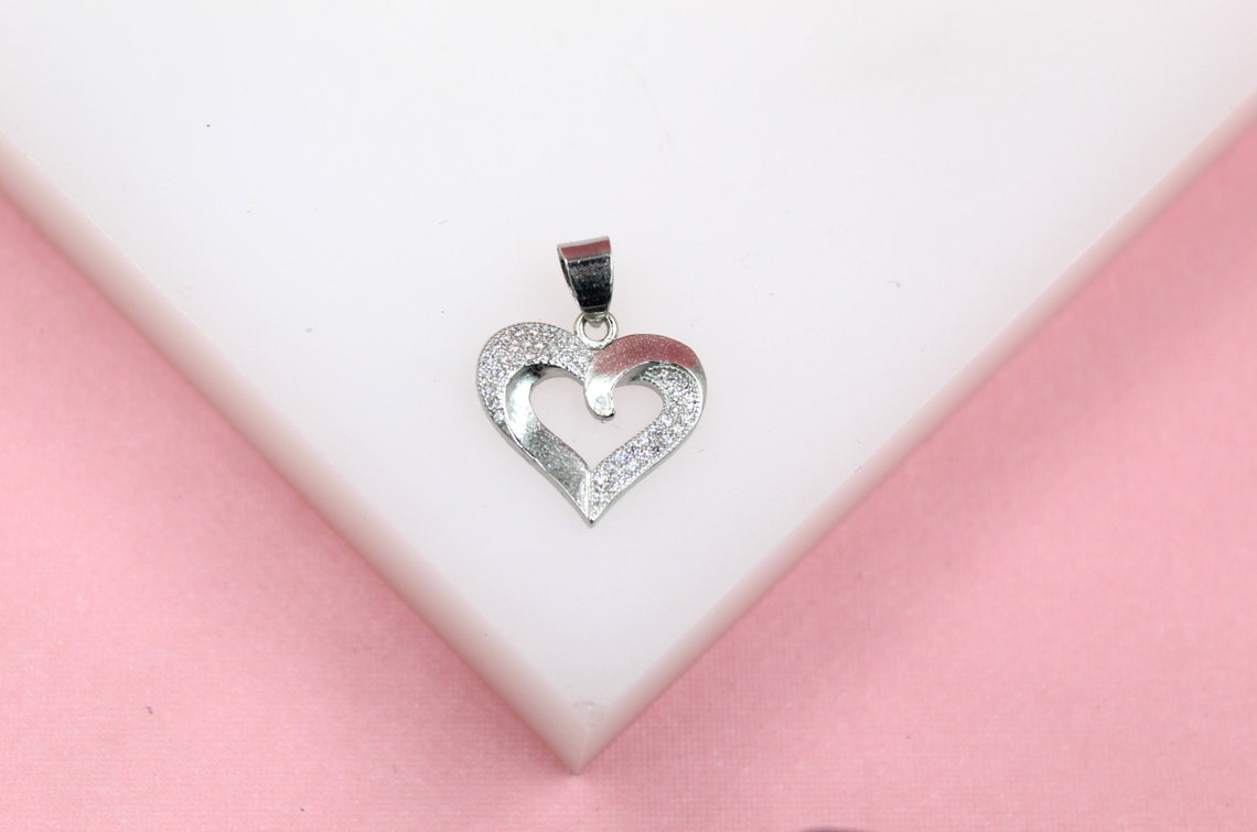 A sparkling silver heart pendant rests on a white display surface against a soft pink background.