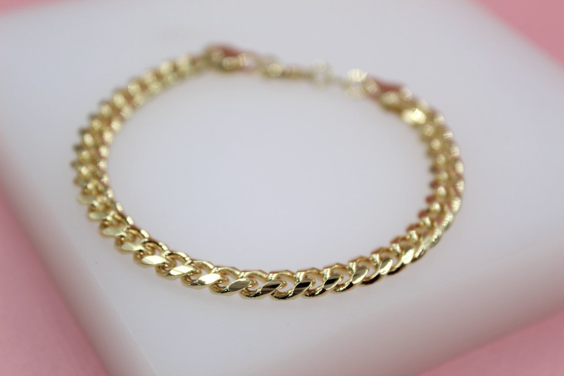 A polished gold curb chain bracelet rests on a clean white surface against a soft pink background.