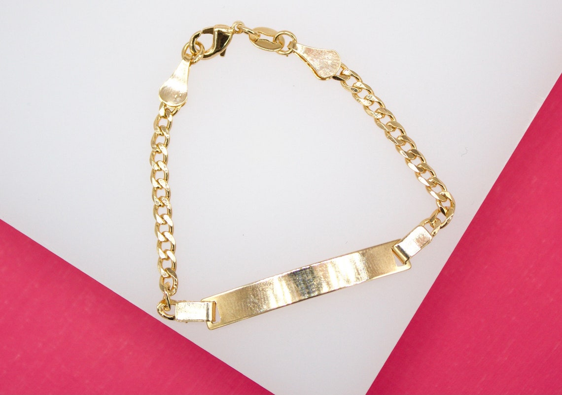 A polished gold ID bracelet with a curb chain rests on a white and pink display surface.