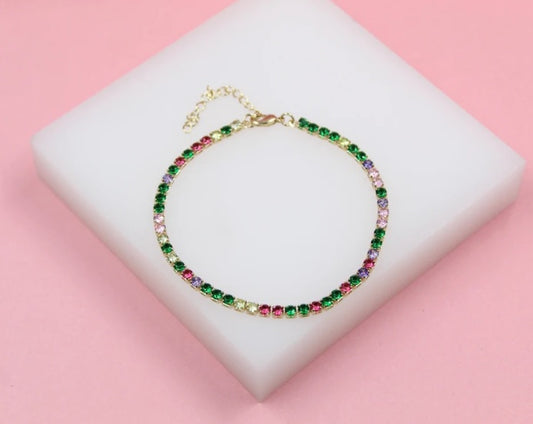 A delicate gold tennis anklet features a colorful gradient of multi-colored crystals on a white display block against a soft pink background.