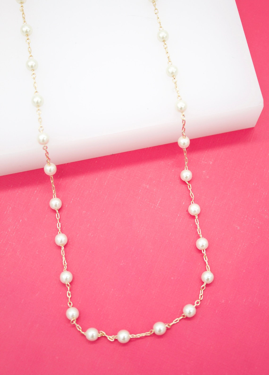 A delicate gold chain necklace with evenly spaced white pearls drapes gracefully over a white surface against a vibrant pink background.
