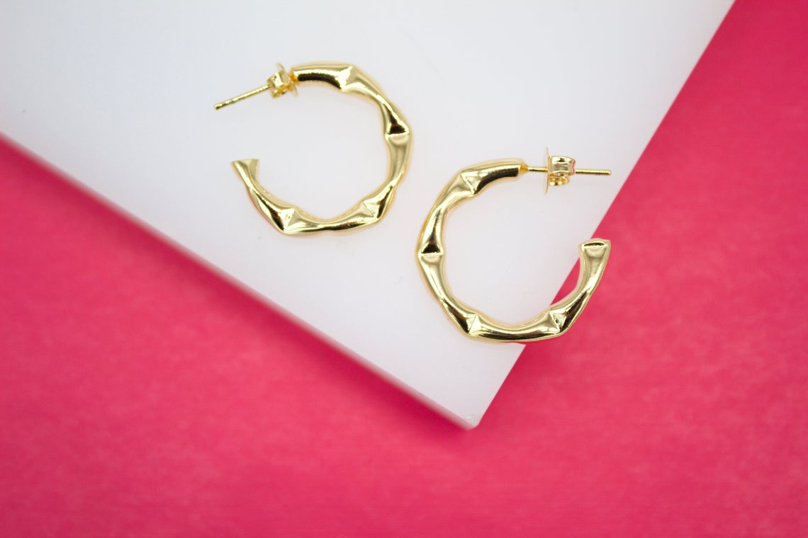A pair of shiny gold bamboo hoop earrings rests on a white display surface against a vibrant pink background.