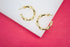 A pair of shiny gold bamboo hoop earrings rests on a white display surface against a vibrant pink background.