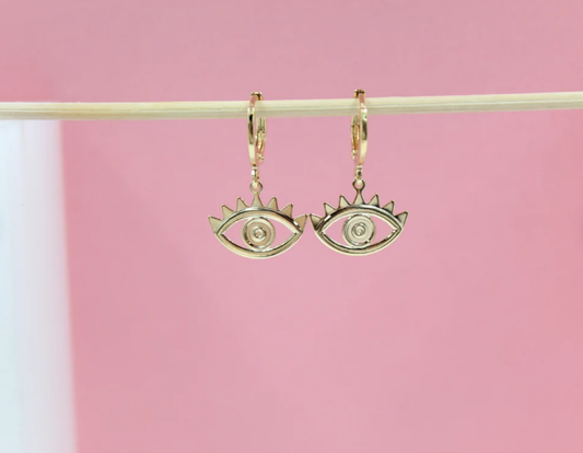 A pair of gold evil eye hoop earrings hangs delicately from a light wooden stick against a soft pink backdrop.