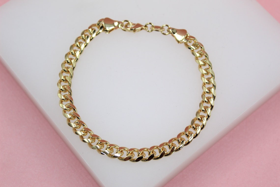 A polished gold cuban link bracelet rests on a white display block against a soft pink background.