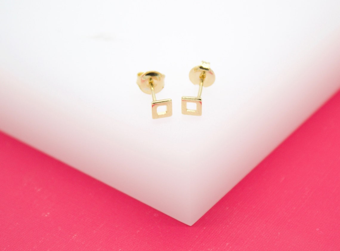 A pair of delicate gold square stud earrings rest neatly on a white and pink display surface.