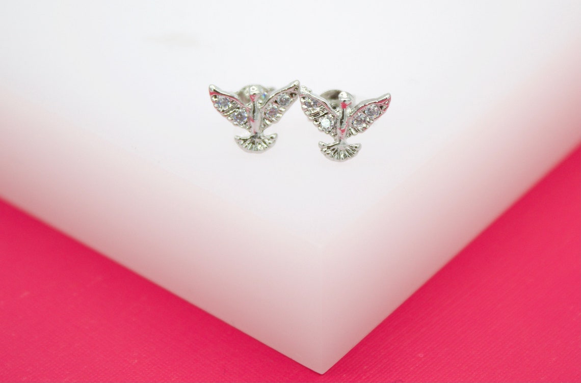 A pair of rhodium-plated Holy Spirit dove stud earrings sparkle with cubic zirconia stones on a white and pink display.
