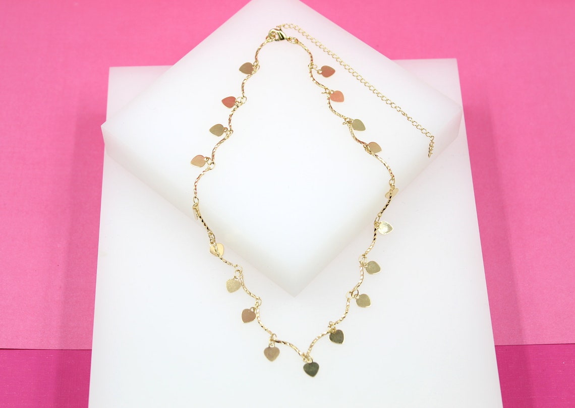 A delicate gold heart-charm necklace rests on a white display block against a vibrant pink background.