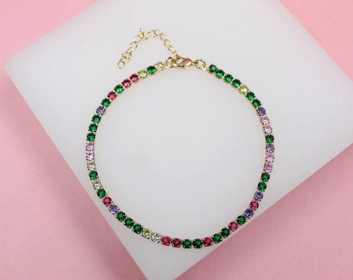 A delicate gold tennis bracelet sparkles with multicolored cubic zirconia crystals in shades of green, pink, and purple against a soft pink background.