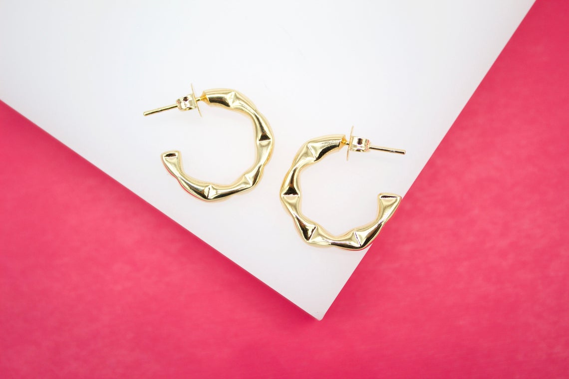 A pair of gleaming gold bamboo-style hoop earrings rests on a white surface against a vibrant pink background.