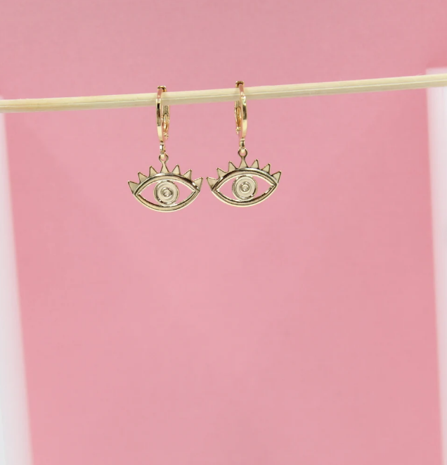 A pair of gold evil eye hoop earrings hangs from a wooden stick against a soft pink background.
