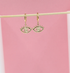 A pair of gold evil eye hoop earrings hangs from a wooden stick against a soft pink background.
