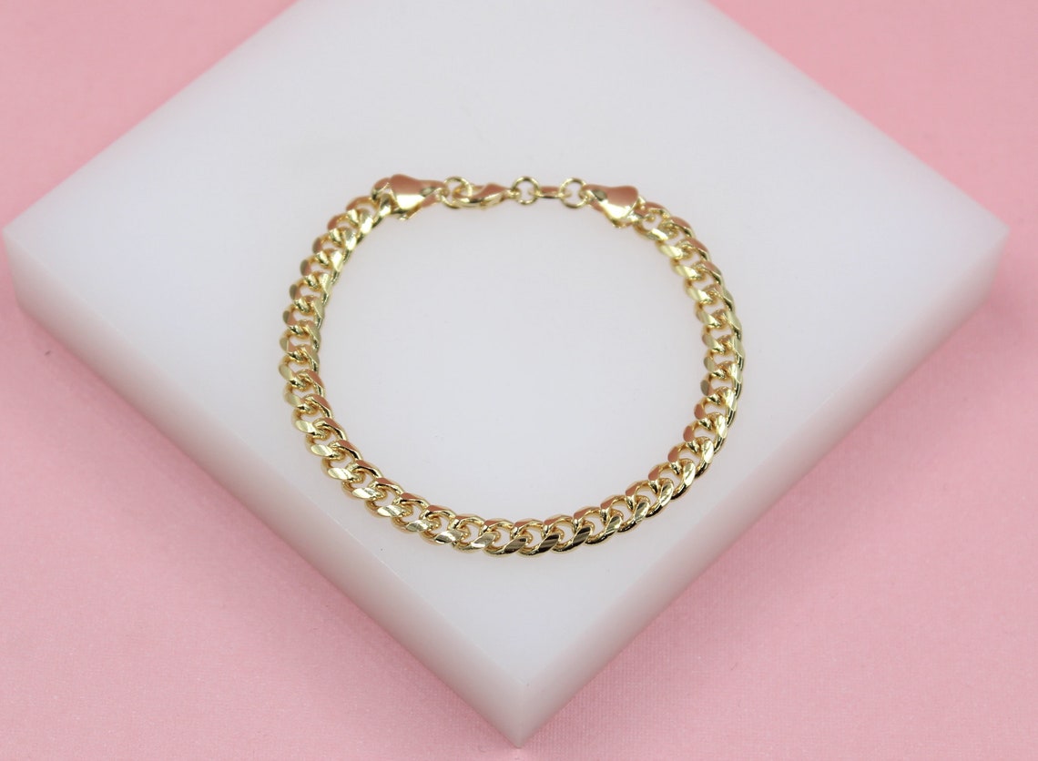 A shiny gold curb chain bracelet rests elegantly on a white display block against a soft pink background.