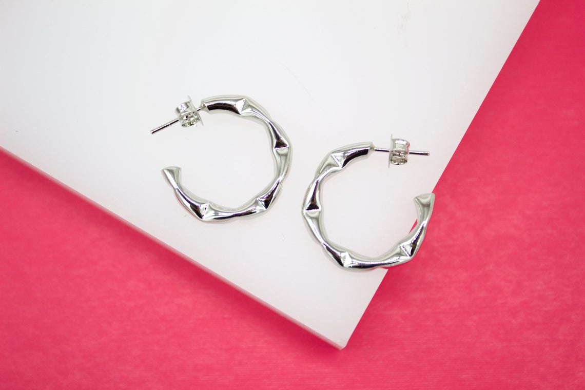 A pair of textured silver hoop earrings rests on a white display against a vibrant pink background.