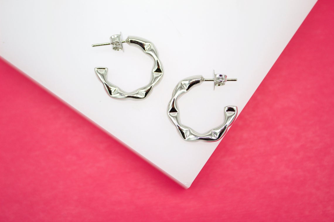 A pair of shiny silver bamboo hoop earrings rests on a white display surface against a vibrant pink background.