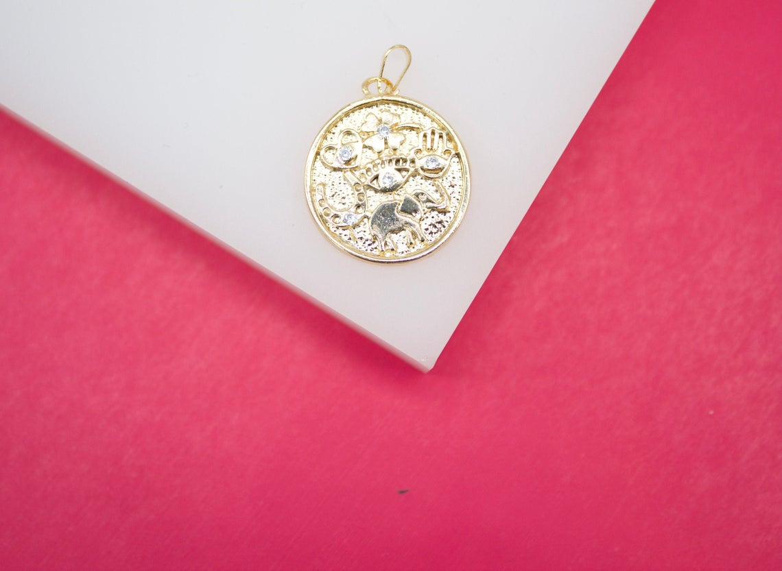 A round gold good luck talisman pendant features engraved symbols accented with tiny cubic zirconia stones against a pink and white background.