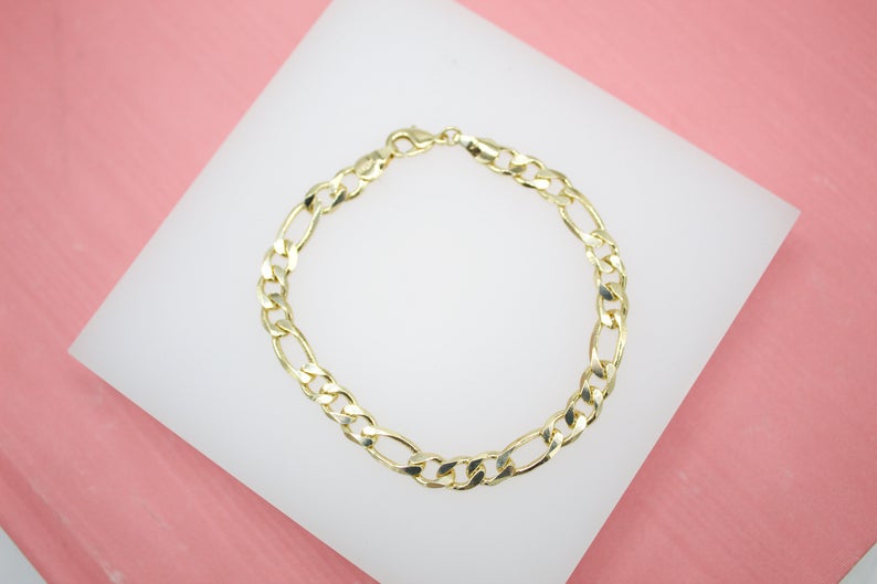 A delicate gold-filled Figaro chain bracelet rests gracefully on a white display against a pink background.