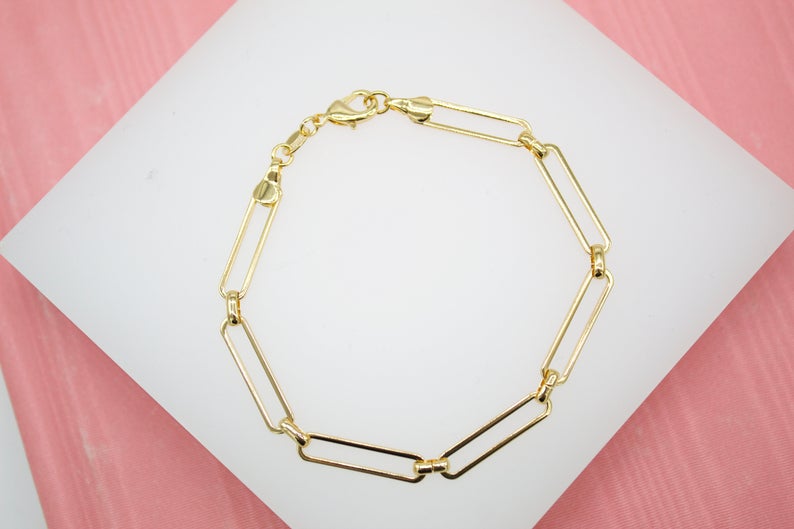 A delicate gold paperclip link bracelet rests on a white display surface against a pink background.