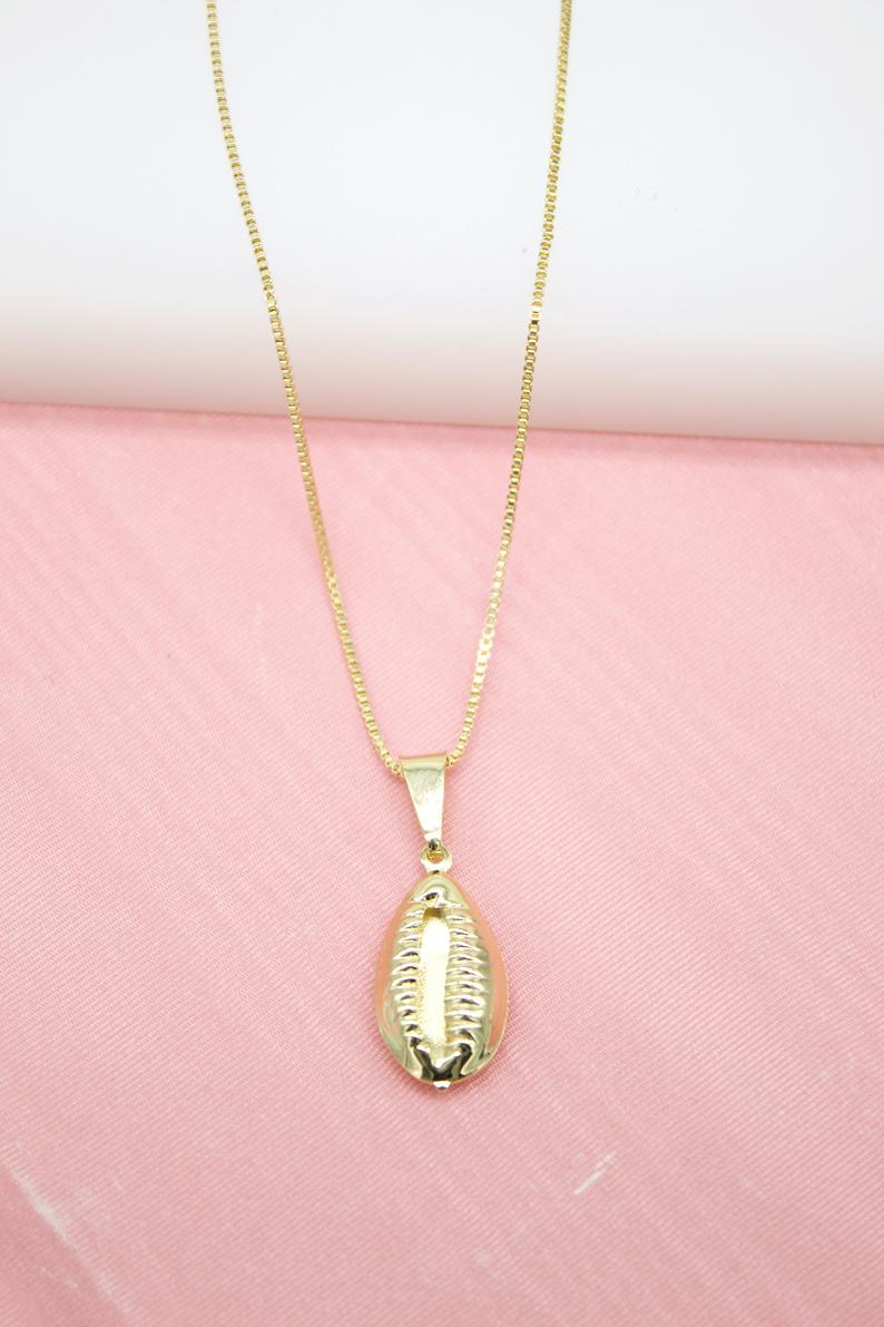 A gold-plated cowrie shell pendant hangs delicately from a matching chain necklace against a soft pink background.
