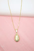 A gold-plated cowrie shell pendant hangs delicately from a matching chain necklace against a soft pink background.