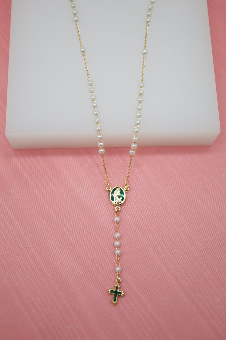 A delicate gold rosary necklace featuring white faux pearls and green enamel accents hangs against a soft pink backdrop.