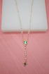 A delicate gold rosary necklace featuring white faux pearls and green enamel accents hangs against a soft pink backdrop.