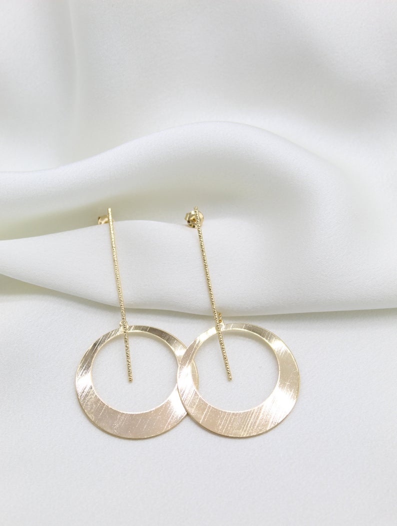 A pair of elegant gold dangle earrings featuring textured circular hoops suspended from delicate post chains rests on a soft white silk background.