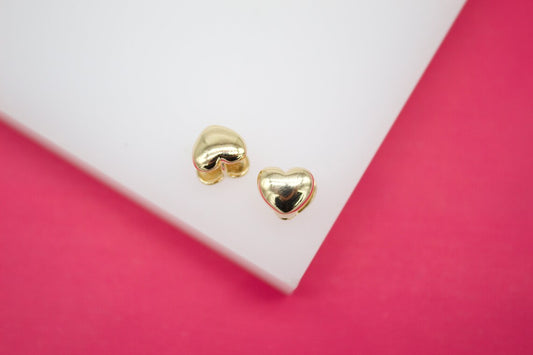 Two shiny gold heart-shaped stud earrings rest on a white display stand against a vibrant pink background.