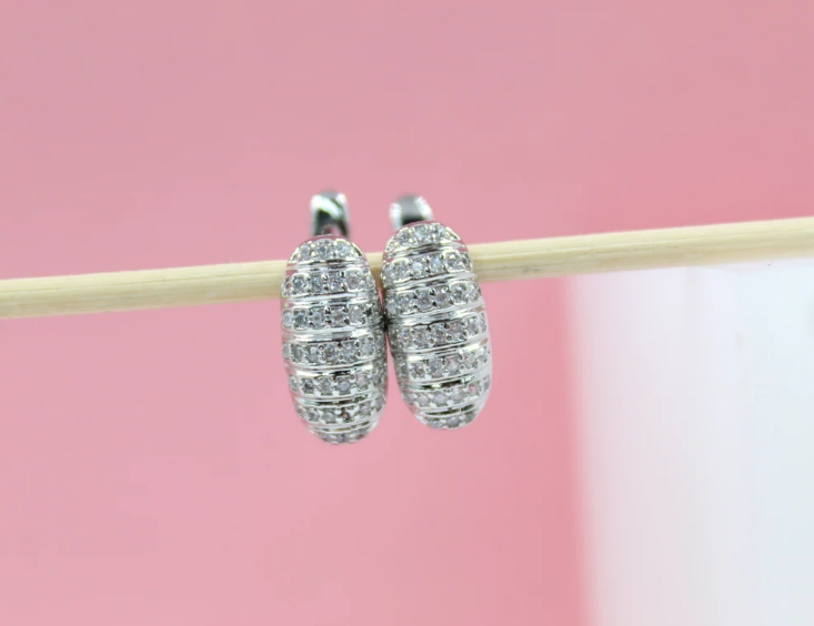 A pair of sparkling pavé-set silver hoop earrings rests on a thin wooden stick against a soft pink background.