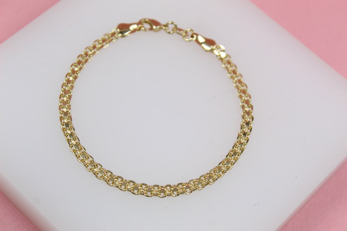 A delicate gold link bracelet rests on a white display stand against a pink background.