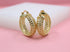 A pair of thick gold hoop earrings with intricate braided link patterns rests on a soft pink and white display block.