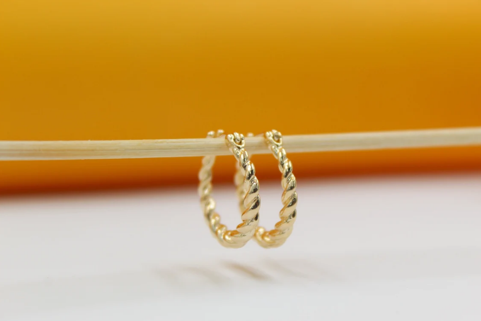 A pair of shiny twisted gold hoop earrings hangs delicately on a wooden stick against a warm yellow and white background.