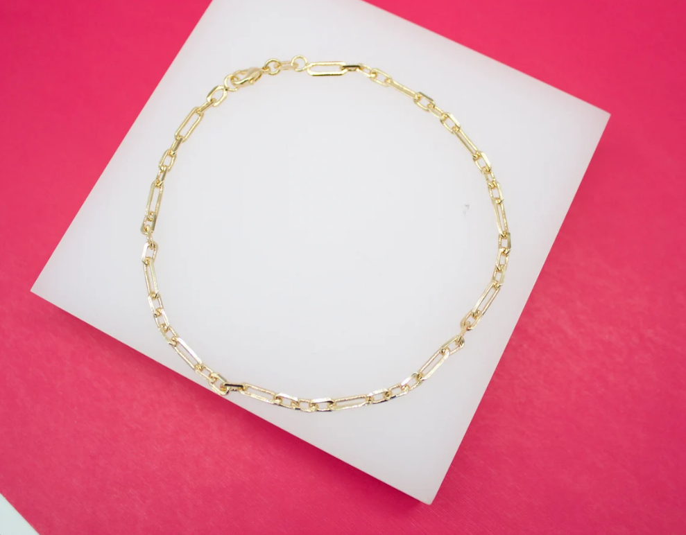 A delicate gold paperclip chain bracelet rests on a white display block against a vibrant pink background.