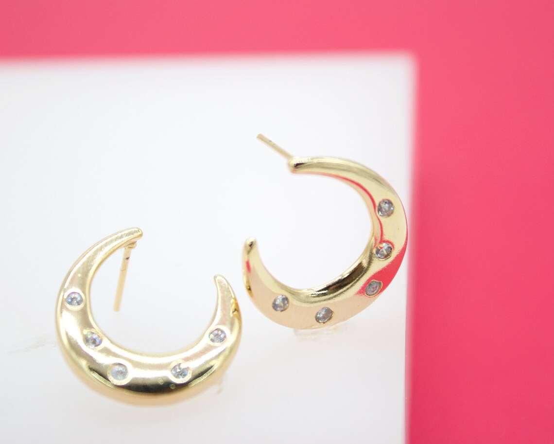 A pair of gold crescent moon hoop earrings embellished with sparkling cubic zirconia stones rests on a white surface against a pink background.