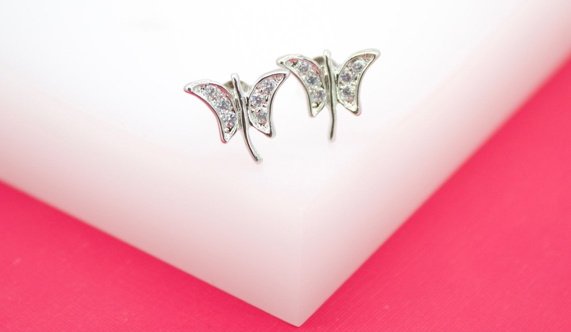 A pair of silver butterfly stud earrings sparkling with cubic zirconia stones rest on a white and pink display.