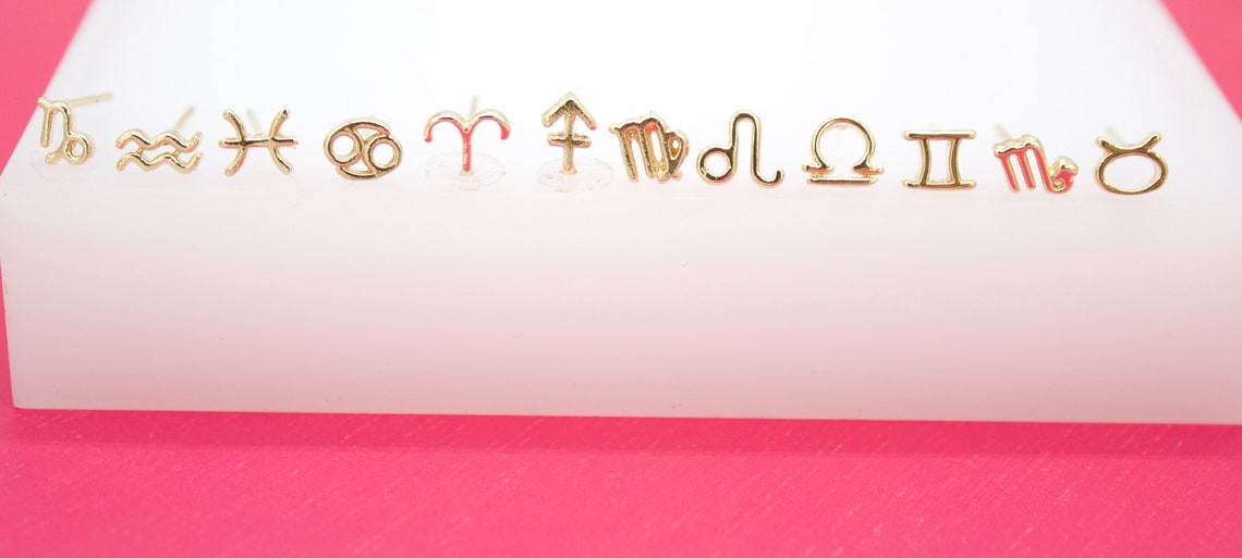 A collection of dainty gold zodiac sign stud earrings lined up on a white display stand against a pink background.