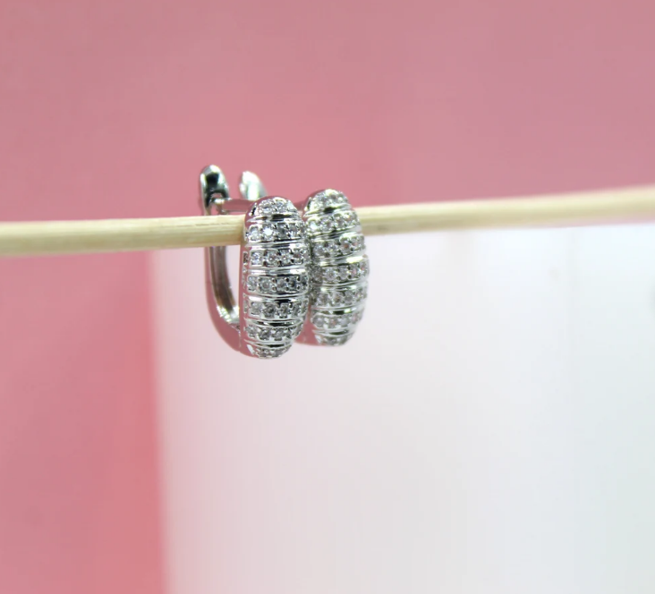 A pair of sparkling silver huggie hoop earrings encrusted with cubic zirconia stones hangs delicately on a wooden stick against a soft pink background.