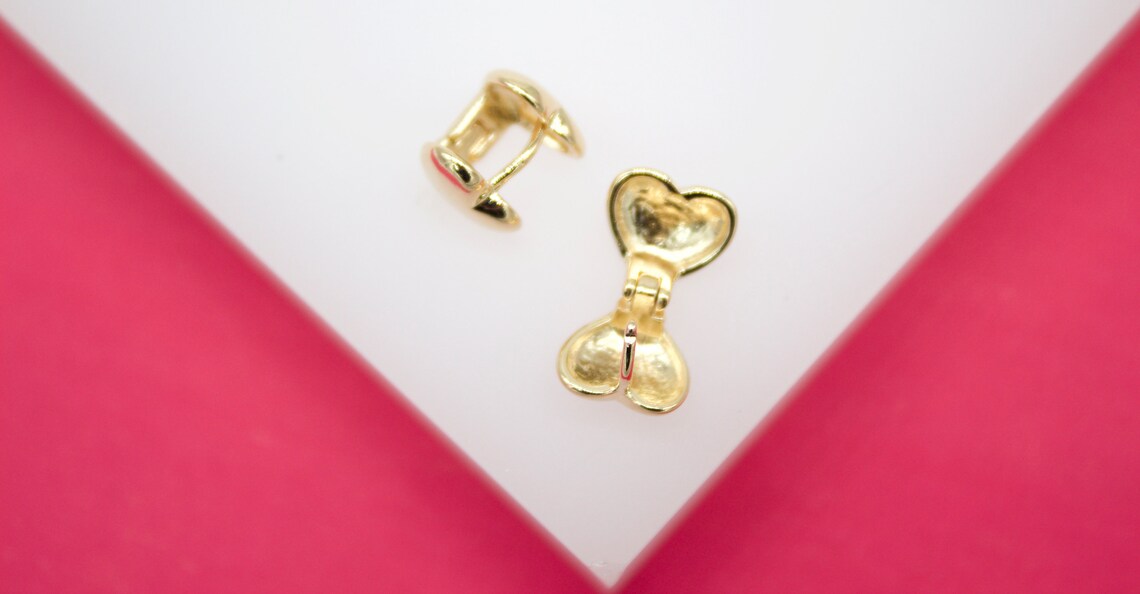 Two gold heart-shaped hoop earrings rest on a white display surface with a pink background.