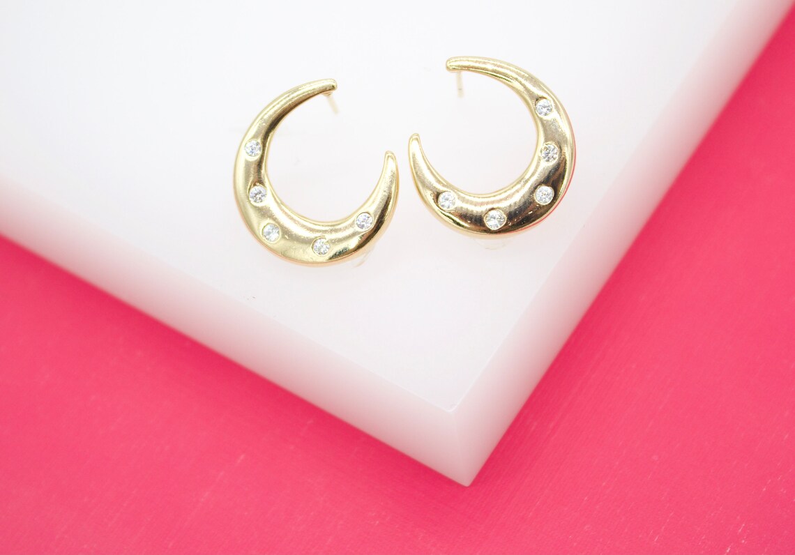 A pair of gold crescent moon hoop earrings studded with cubic zirconia stones rests on a white display surface against a vibrant pink background.