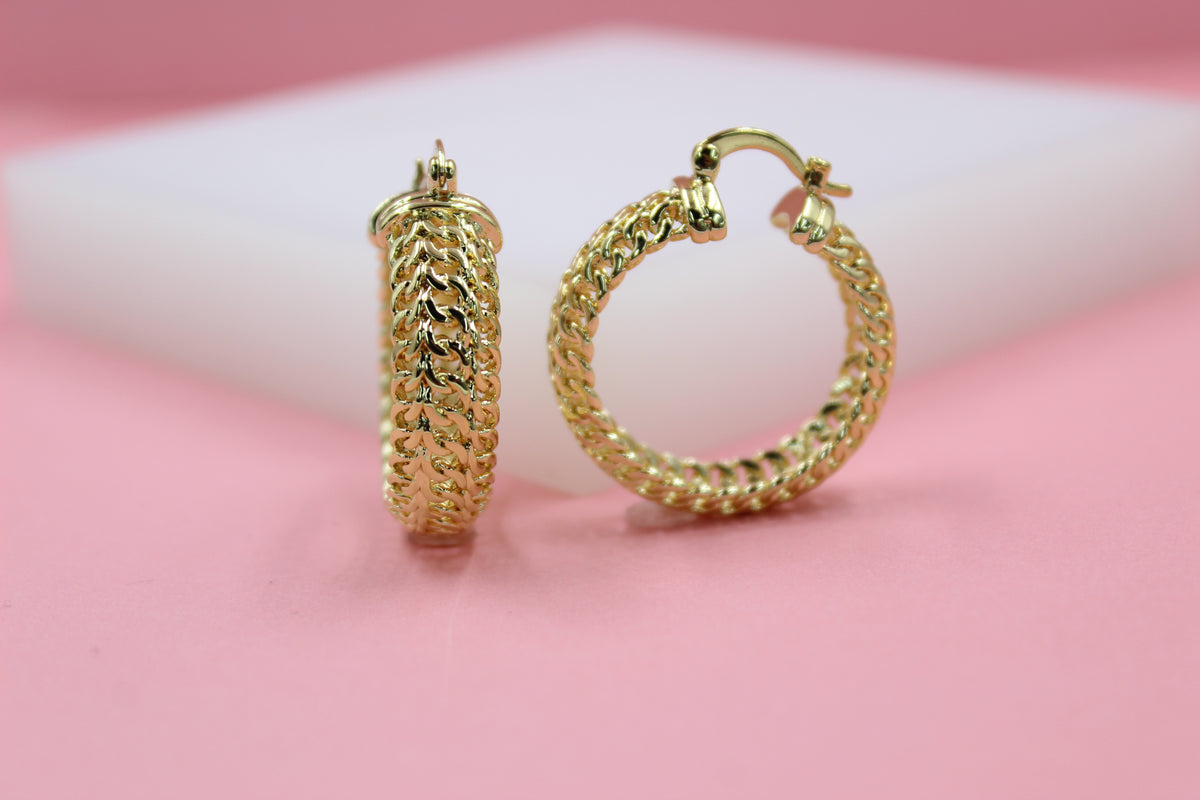 Two shiny gold hoop earrings with an intricate chain-link design rest on a pink surface.