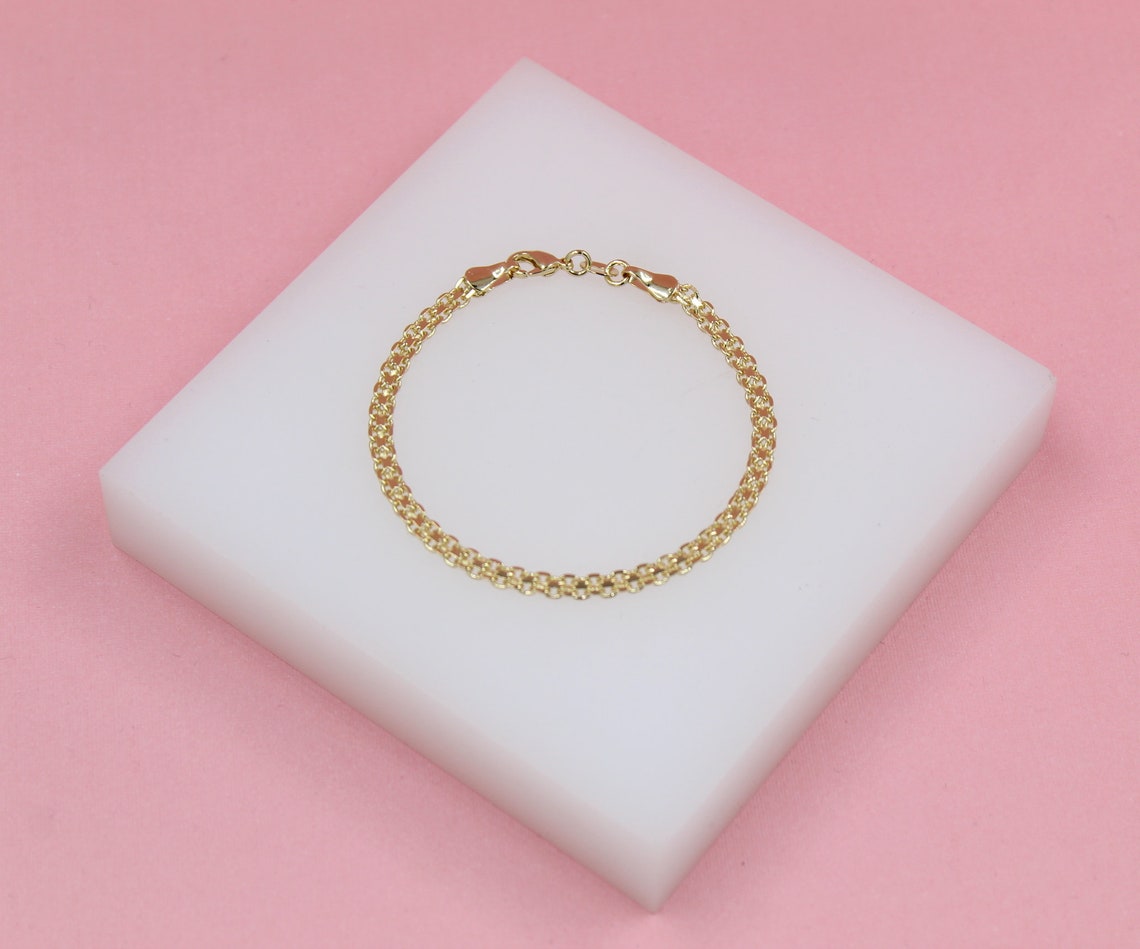A delicate gold chain bracelet rests centered on a white display block against a soft pink background.
