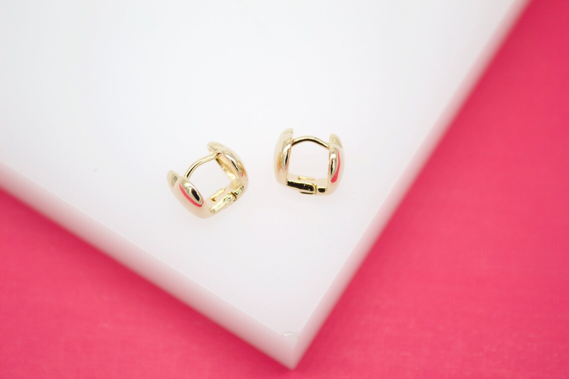 A pair of small gold huggie hoop earrings rests on a white and pink display surface.