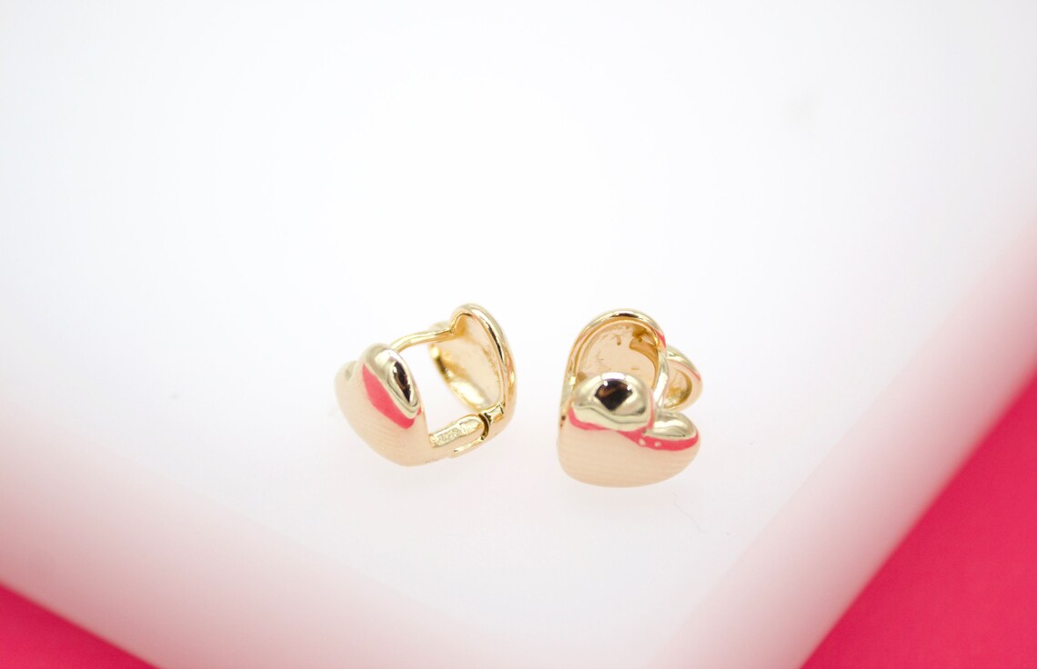 A pair of shiny gold heart-shaped huggie earrings rests on a white display surface.