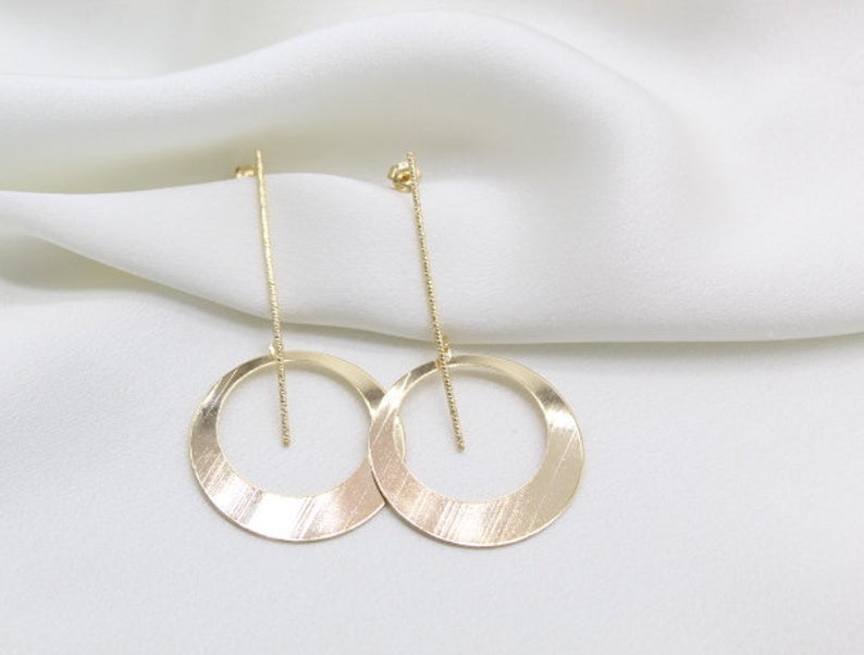 A pair of minimalist gold drop earrings featuring slender textured bars and dangling flat hoops rests elegantly on a soft white silk background.