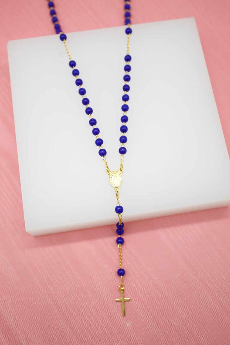 A Catholic rosary necklace featuring vibrant blue beads and a gold chain, centerpiece, and crucifix lies displayed on a white surface against a pink background.