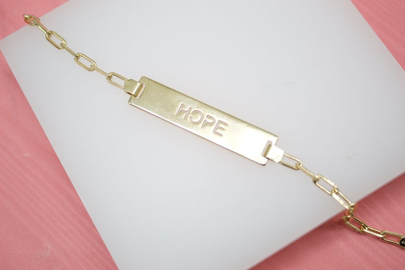 A gold ID bracelet rests on a white display block, showcasing an engraved rectangular plate with the word "HOPE."