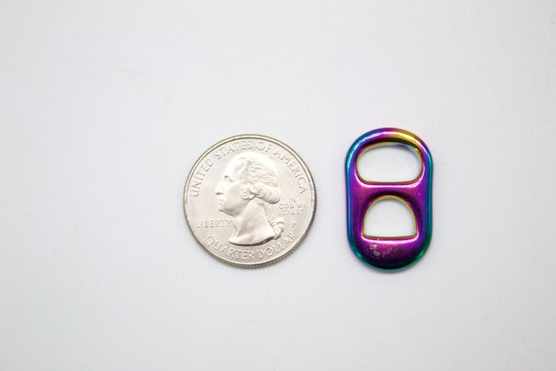 A vibrant rainbow-colored soda tab sits beside a United States quarter for scale on a neutral background.
